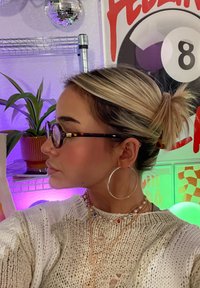 Προφίλ ενός ατόμου με ξανθά μαλλιά σε κότσο, φορώντας γυαλιά με καβουρίσια κορν�ίζα, ασημένια κρίκους σκουλαρίκια και στρωματοποιημένα κολιέ, μπροστά σε πολύχρωμο φόντο.