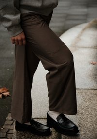 Hnedé nohavice širokého strihu z textúrovaného materiálu, s vyšívaným detailom "Zakázané" na stehne, doplnené lesklými čiernymi mokasínami.