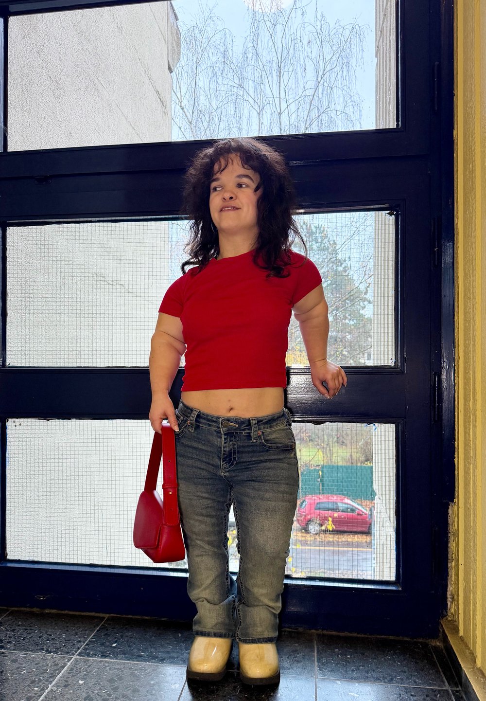 Haut rouge ajusté à manches courtes, jean en denim bleu, chaussures compensées jaunes, sac à main rouge, debout près d'une grande fenêtre avec lumière naturelle.