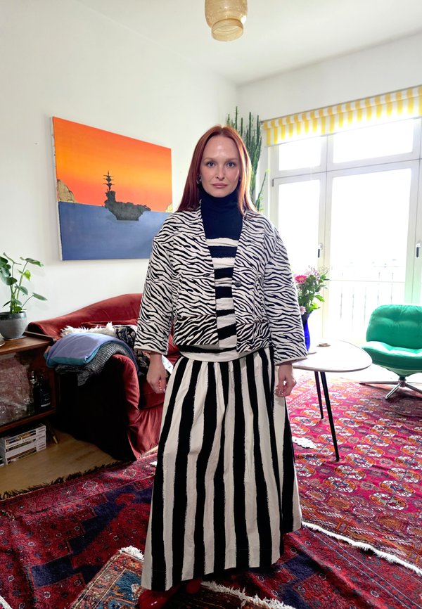 Femme aux cheveux roux portant une veste à imprimé zèbre et une jupe à rayures noires et blanches, debout dans un salon coloré avec des plantes et des œuvres d'art.