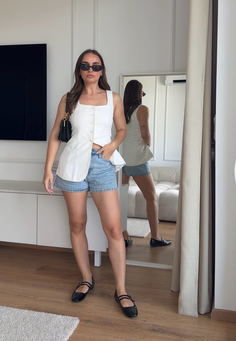 Femme portant un haut blanc sans manches boutonné, un short en denim, des ballerines noires et des lunettes de soleil, debout près d'un miroir dans un salon moderne.