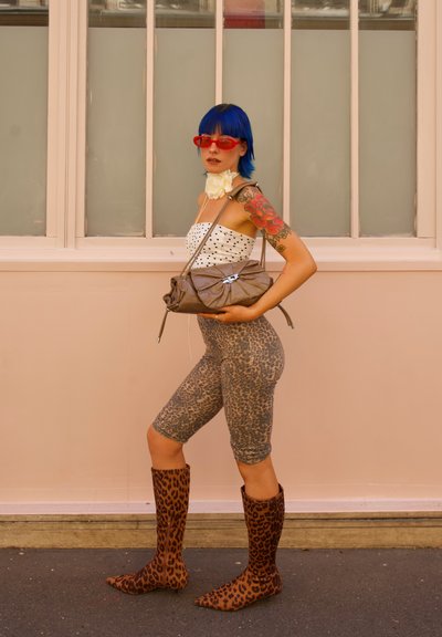 Leggings con estampado de leopardo, blusa de lunares, bolso marrón con lazo y botas altas de estampado de leopardo. Cabello azul, gafas de sol rojas y redondas, acento floral.