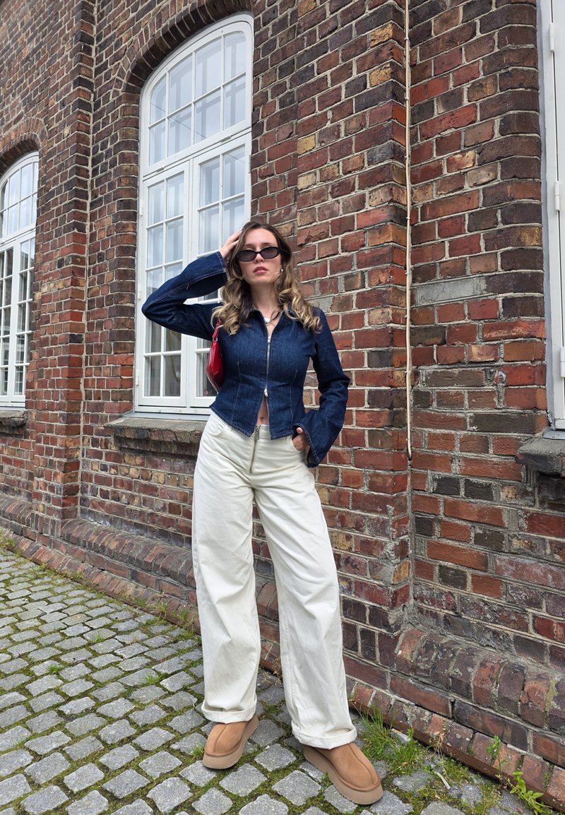 Veste en denim au design ajusté et aux manches évasées, associée à un large pantalon blanc et des souliers à plateforme marron. Sac à main rouge en accent.