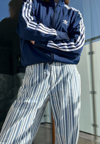 Jacket de fecho éclair azul-marinho da Adidas com riscas brancas, emparelhado com calças riscas azuis e brancas. Tecido suave com um corte relaxado.