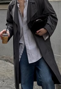 Langer grauer Trenchcoat über einem gestreiften Hemd mit Knöpfen, blaue Jeans, hält eine Kaffeetasse und eine schwarze Clutch unter dem Arm.