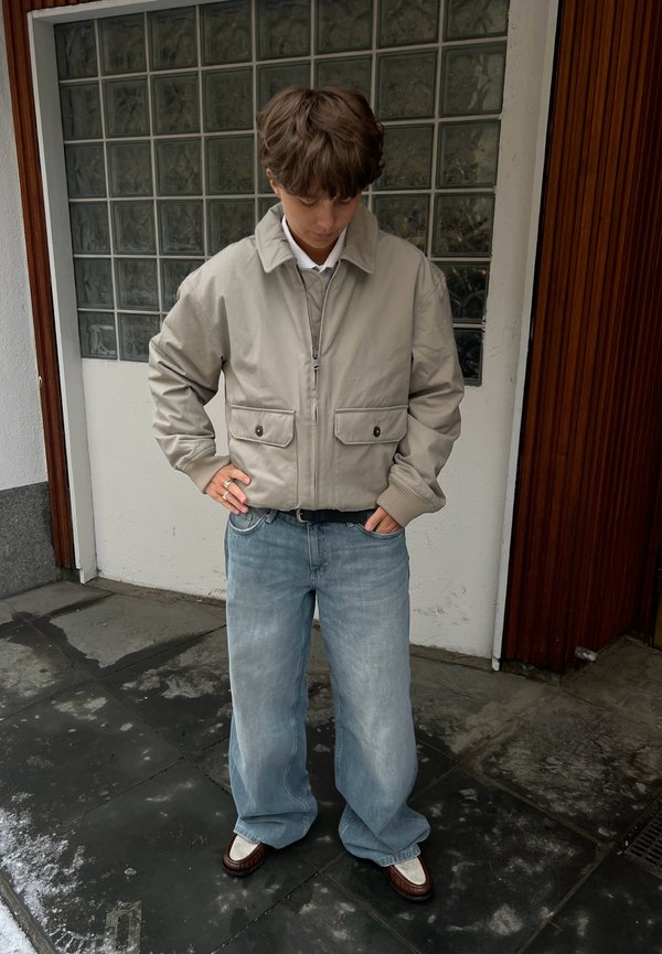 Ung person iført beige jakke, lyseblå vide jeans og mørkebrune sko, står med hendene i lommene på våt fortau.