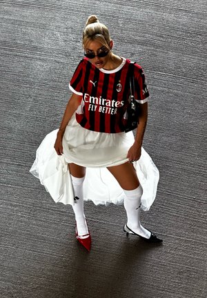 Raudonai ir juodai dryžuota futbolo marškinėliai, baltos sijonas su tiuliu, baltos kelio ilgio kojinės, juodi ir raudoni aukštakulniai, švelniai surišti plaukai į uodegėlę.