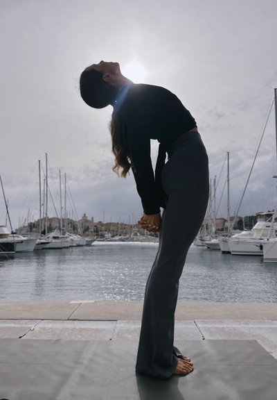 Top negro de manga larga y pantalones ajustados grises. Persona en una pose de flexión, con un fondo de waterfront que presenta barcos y un cielo nublado.