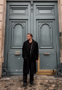 En man i en svart ullkappa står vid utsmyckade ljusblå dubbeldörrar med dekorativa paneler och ett guldhandtag. Stengolvet är belagt med kullerstenar.