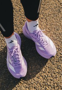 Lichtpaarse hardloopschoenen met witte accenten, een geribbelde mesh bovenkant en platte veters, gecombineerd met witte sokken met een zwart logo.