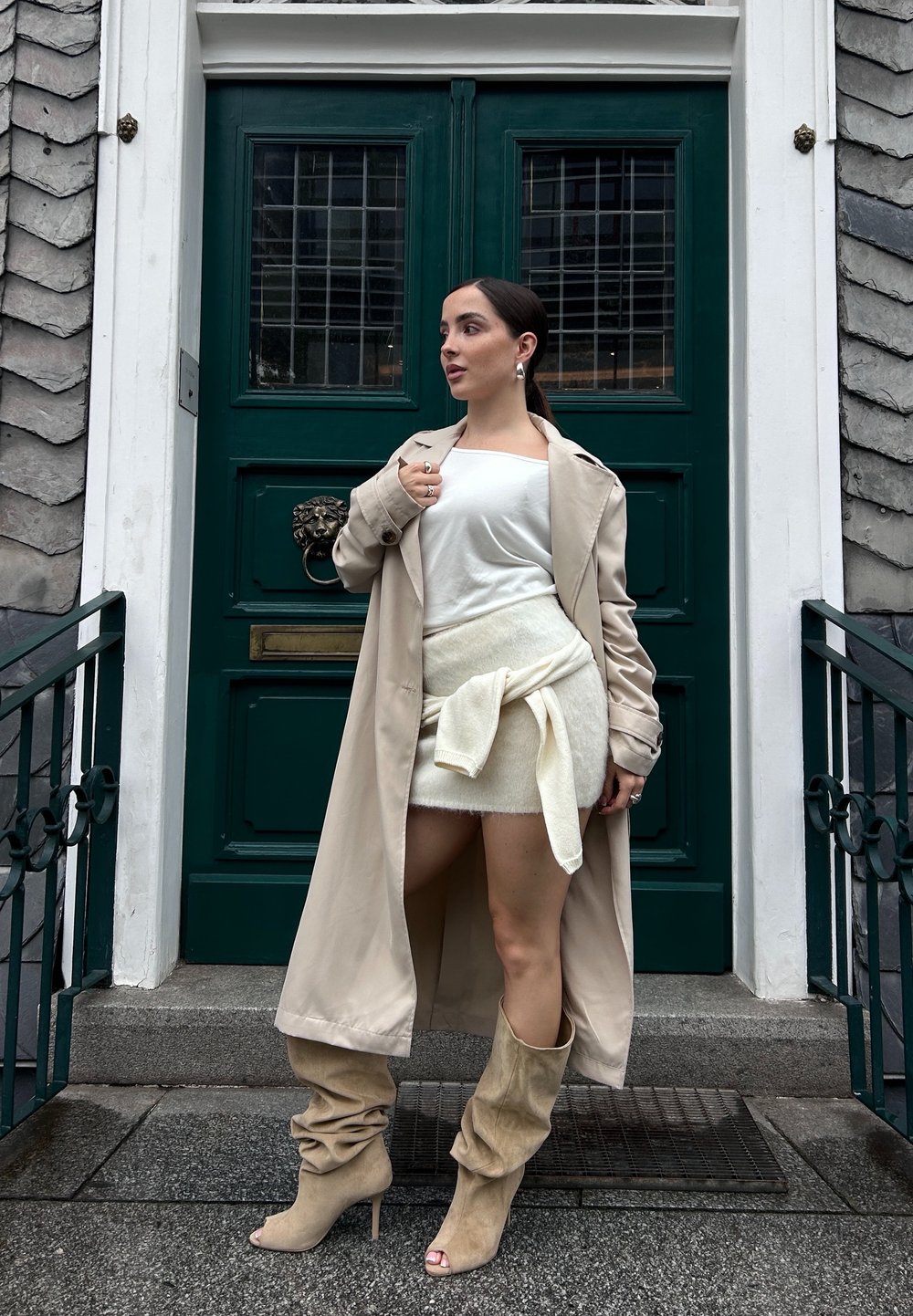 Beige trenchcoat, hvid top, lys sweaternederdel med bånd, og beige slouchy højhælede støvler. Står ved en grøn dør med en løvehoveddørklokke.