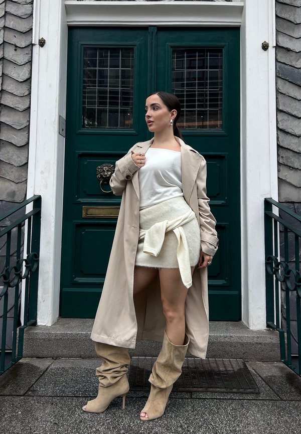 Beige trenchcoat, witte top, lichte sweaterrok met strik, en beige loszittende hoge hakkenlaarzen. Staande bij een groene deur met een leeuwenkopklopper.