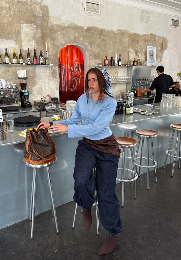 Žena u plavoj majici i marami na glavi sjedi uz metalni šank s barskim stolicama, seže u smeđu dizajnersku torbu u modernom kafiću.