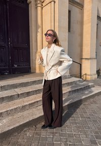 Nadrozmerné krémové sako s opaskom cez biely top, doplnené širokými hnedými nohavicami a čiernymi topánkami, na pozadí kamenných schodov.