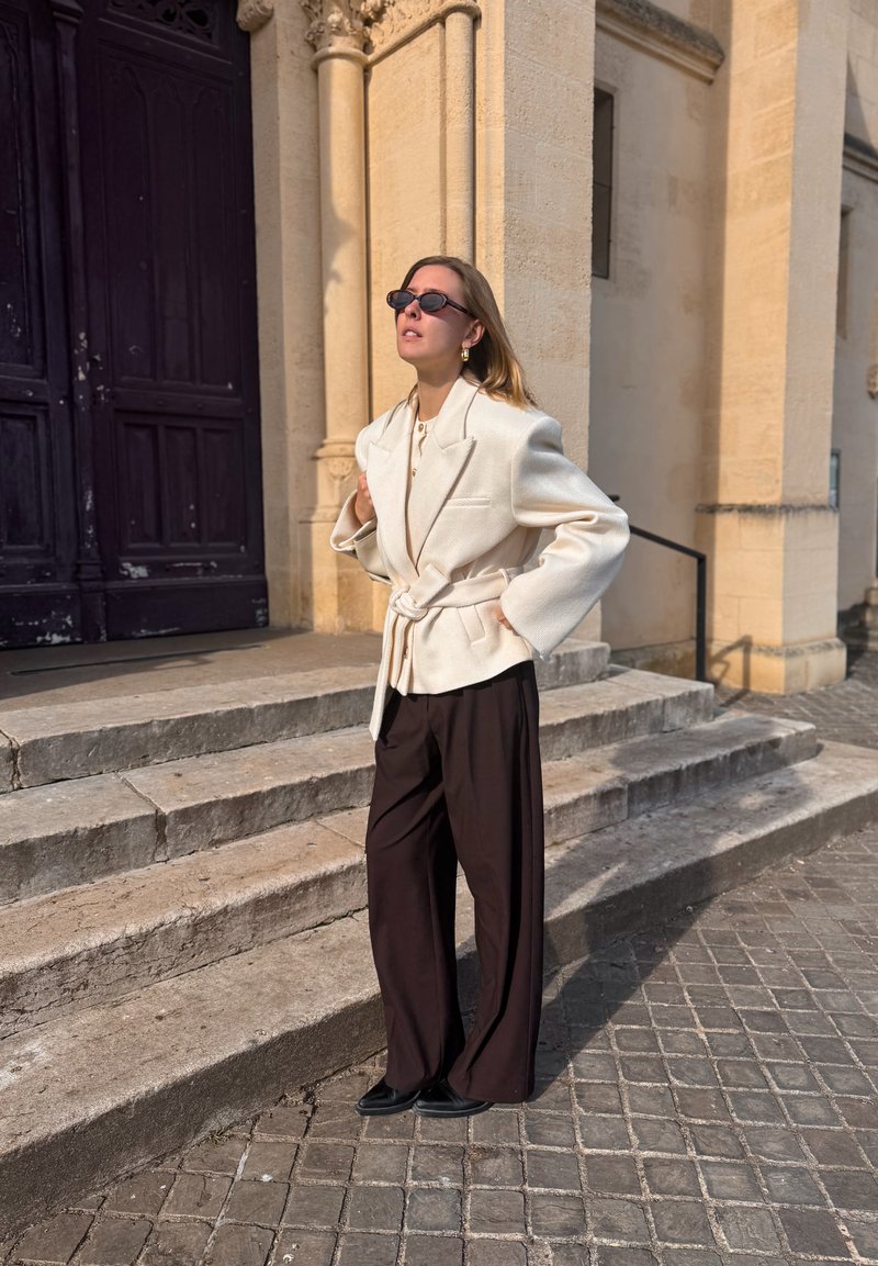 Blazer crem supradimensionat cu curea peste un top alb, asortat cu pantaloni maro cu picioare largi și pantofi negri, pe fundalul unor trepte din piatră.