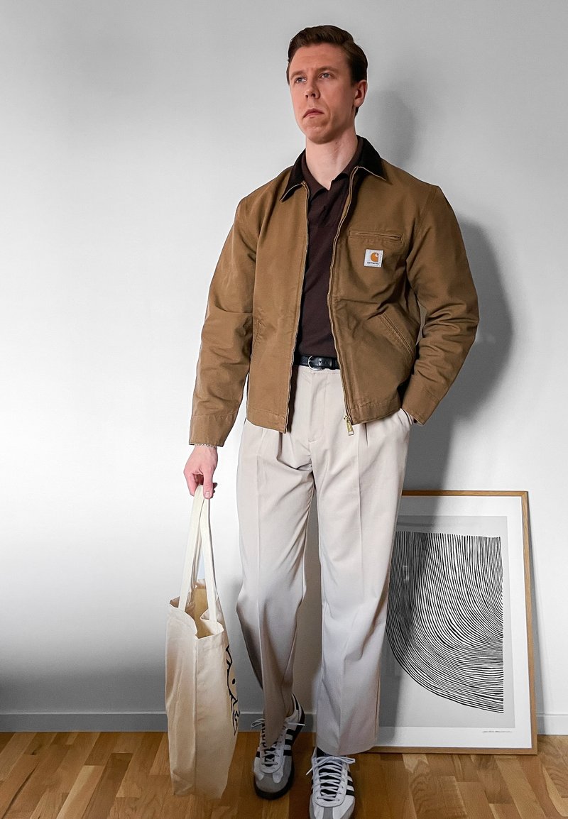 Bărbat purtând jachetă maro, cămașă neagră, pantaloni bej și adidași, ținând o geantă de pânză, stând lângă o lucrare de artă alb-negru încadrată pe podea.