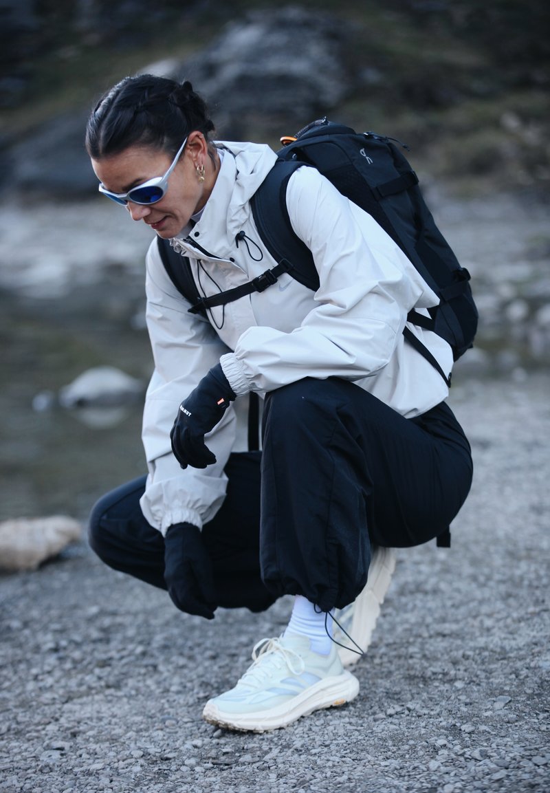 Woman wearing sunglasses, white jacket, black pants, gloves, and backpack crouches on rocky ground near water.