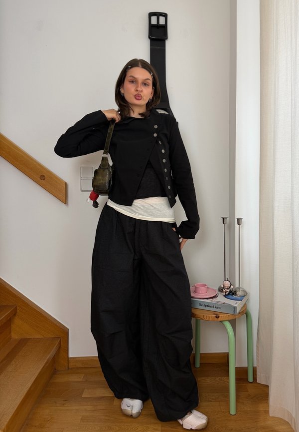 Joven mujer con chaqueta negra y pantalones anchos posa con pequeño bolso de hombro cerca de unas escaleras y una pequeña mesa con libros y lámpara.