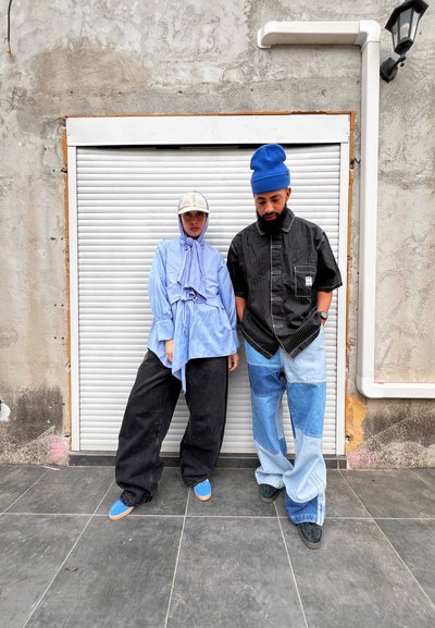 Una persona lleva una camiseta de rayas azules de manga larga y pantalones anchos negros, mientras que la otra lleva una camisa negra y jeans de patchwork, ambas con sombreros, de pie contra una pared.