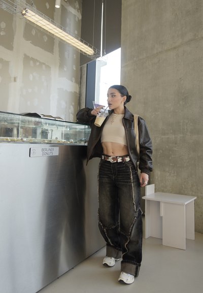 Chaqueta de cuero marrón, top corto beige, jeans de mezclilla negra desgastados con flecos, zapatillas blancas, vaso transparente con bebida, interior moderno.