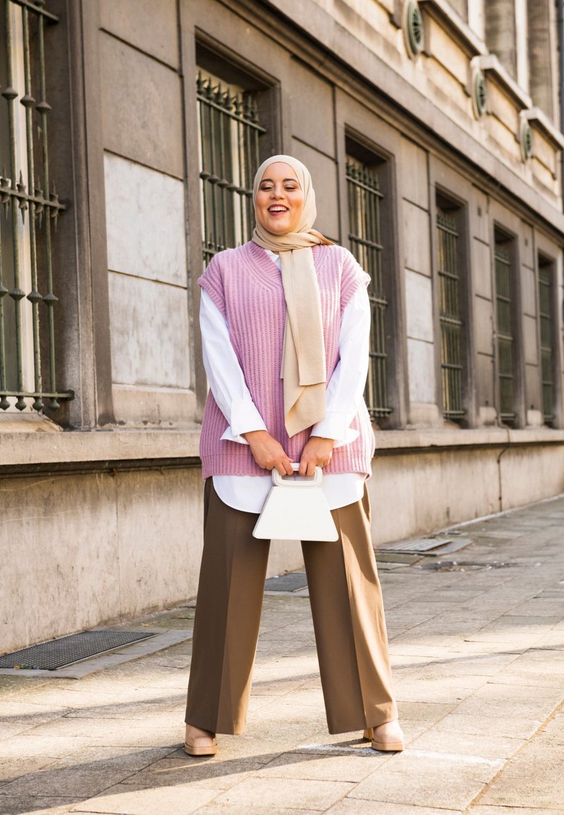 Light pink knitted vest over a white shirt, paired with brown wide-leg trousers. Features a white geometric handbag and beige shoes.