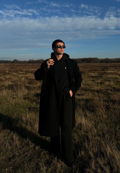 Abrigo negro sobre una sudadera negra con capucha, pantalones amplios negros y gafas de sol oscuras, con un gorro, de pie en un campo de hierba bajo un cielo azul.