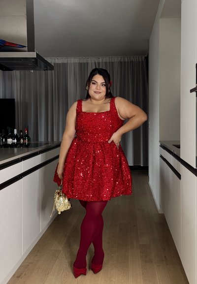 Vestido rojo de lentejuelas con escote cuadrado y falda acampanada. Combinado con medias burdeos y tacones rojos de punta, sosteniendo un bolso dorado.