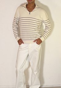 Homme portant un pull à fermeture éclair crème avec de fines rayures horizontales brunes et un pantalon blanc, debout les mains dans les poches contre un mur uni.