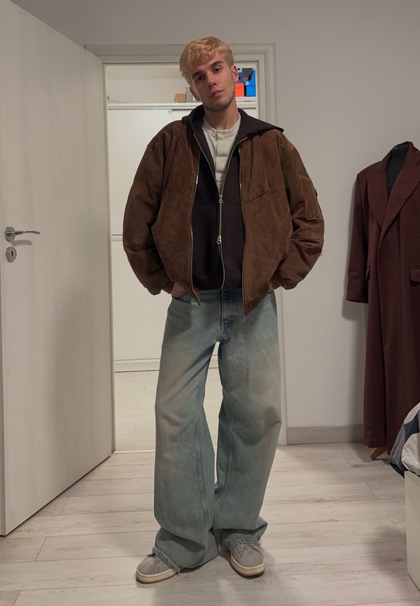 Brązowa zamszowa, oversize'owa kurtka na ciemnym hoodie, zestawiona z jasnymi, wyblakłymi, szerokimi spodniami i szarymi trampkami. Proste tło z drzwiami.