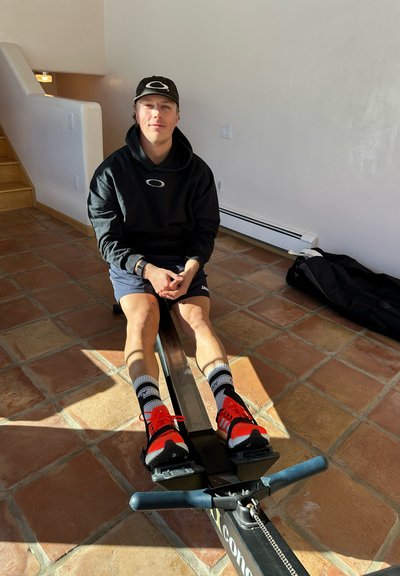 Máquina de remo con un riel de metal elegante y soportes para los pies. El usuario lleva puesta una sudadera negra, pantalones cortos, zapatos naranjas y calcetines a rayas. Entorno interior iluminado por el sol.