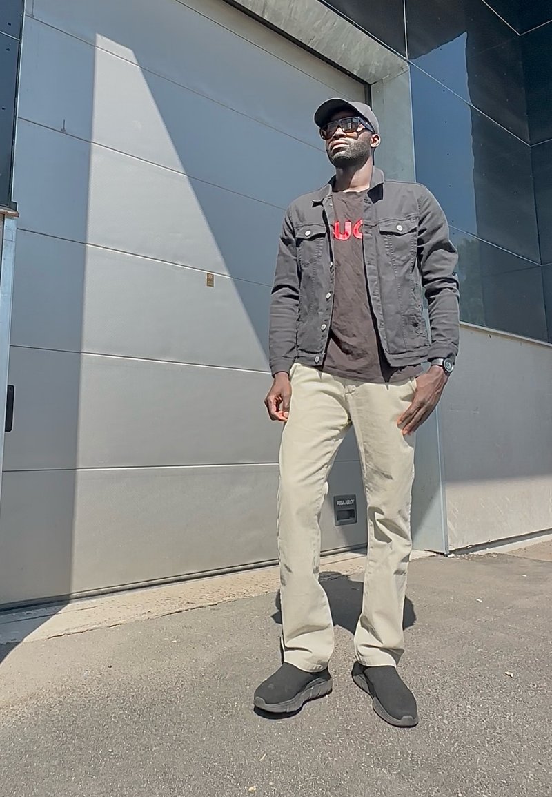 Homme portant une veste noire, une casquette noire, des lunettes de soleil, un pantalon beige et des chaussures noires, debout à l'extérieur près d'un bâtiment moderne en plein soleil.