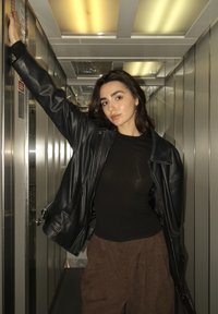 Jauna sieviete melnā ādas jakā un brūnās biksēs, pozējot ar vienu roku paceltu metāliskā liftā ar fluorescējošiem griestu apgaismojumiem.