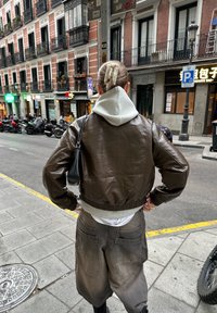 Brun læder cropped jakke med elastisk talje over en grå hættetrøje. Mørk taupe brede bukser, minimalistisk sort håndtaske.