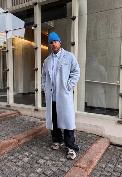 Hombre de pie sobre una acera adoquinada, vestido con un abrigo largo azul claro, gorro de punto azul, pantalones negros y zapatillas deportivas, cerca de un edificio de cristal.