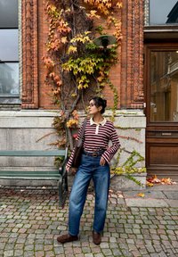 Gestreept top met lange mouwen en een lichte kraag, blauwe jeans met wijde pijpen, bruine mocassins en een bruine schoudertas. Achtergrond van groene en rode klimop.