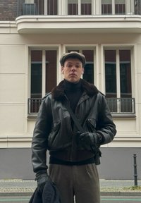 Veste bomber en cuir noir avec col en fourrure, col roulé, pantalon gris et casquette plate ; tenant un manteau sombre. Fond urbain avec des fenêtres.