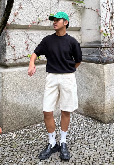 Top corto de punto negro con textura, pantalones bermuda de cuadros crema, gorra de béisbol verde, mocasines negros, calcetines blancos con logo, fondo de piedra.