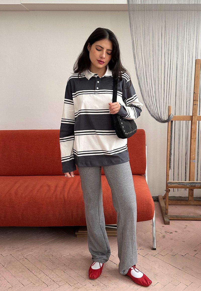 Jeune femme portant un t-shirt à manches longues à rayures noires et blanches, pantalon gris, ballerines rouges, tenant un sac à main noir, debout devant un canapé orange.