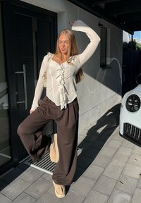 Camicia bianca con volant sul davanti, abbinata a pantaloni ampi marrone scuro. Scarpe beige completano il look, con uno sfondo di architettura moderna.