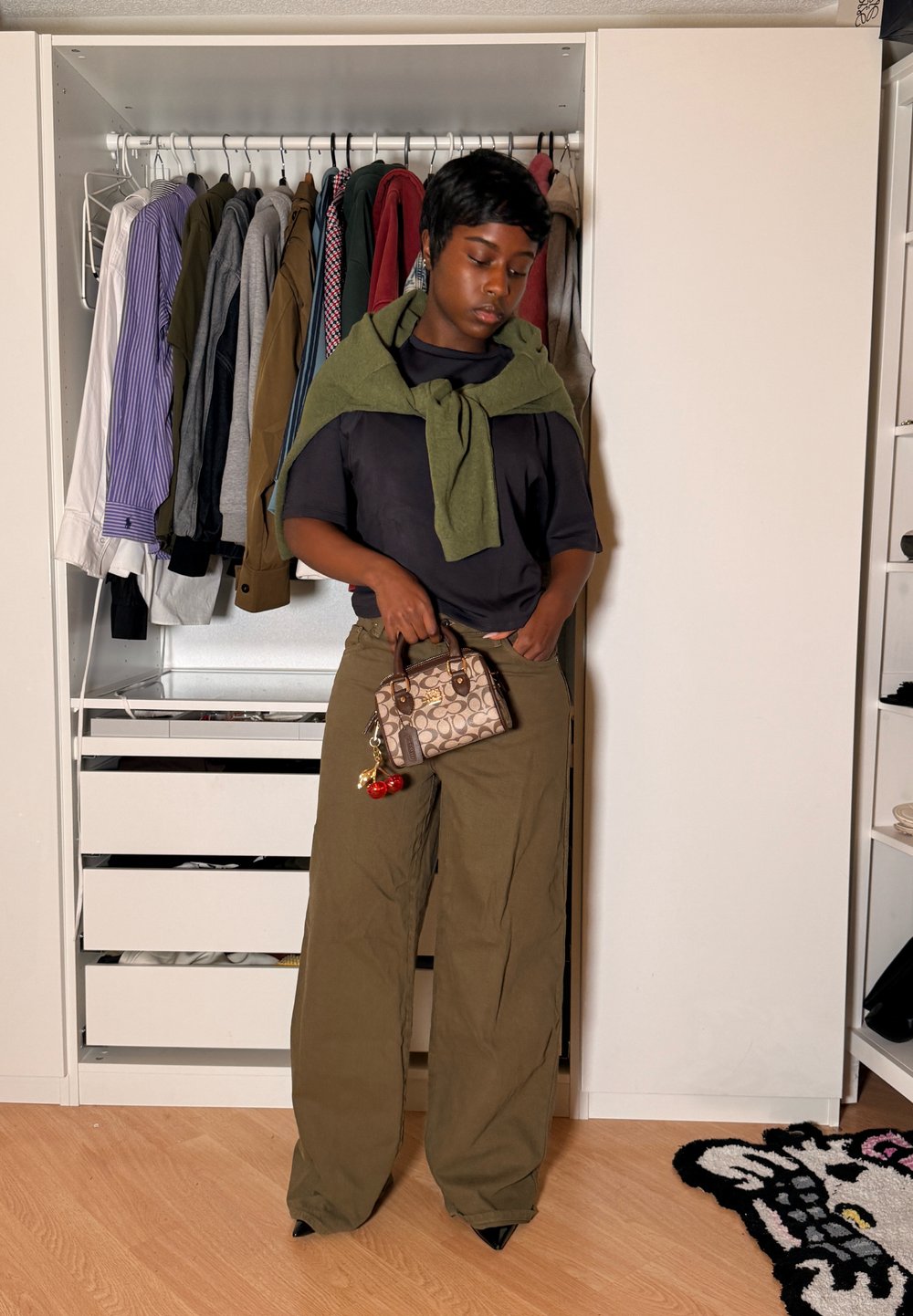 Personne debout devant une armoire ouverte, portant une chemise noire, un pantalon olive, un pull vert drapé sur les épaules, tenant un sac à main à motifs.