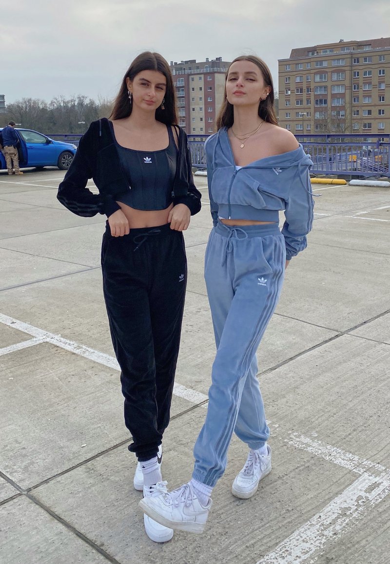 To modeller bærer matchende afslappede træningsdragter; den ene i sort velour og den anden i lyseblå, begge med cropped tops og hvide sneakers.