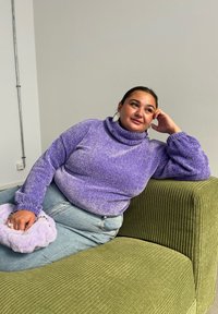 Lilla, glitrende turtleneck genser med lange ermer, kombinert med lyseblå jeans og en myk lavendelveske, sittende på en grønn kordfløyelssofa.
