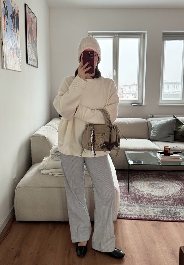 Maglione crema oversize con polsini a costine, pantaloni a righe a gamba larga, berretto rosa lavorato a maglia e borsa a tracolla beige con dettagli metallici.
