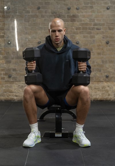 Unas pesas negras sostenidas en posición sentada por una persona que lleva una sudadera oscura y pantalones cortos sobre un suelo de gimnasio negro, con un fondo de pared de ladrillo.