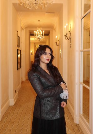 Blazer texturizado en negro y plata con un cinturón en la cintura, combinado con una falda negra transparente. Situado en un pasillo bien iluminado con candelabros.