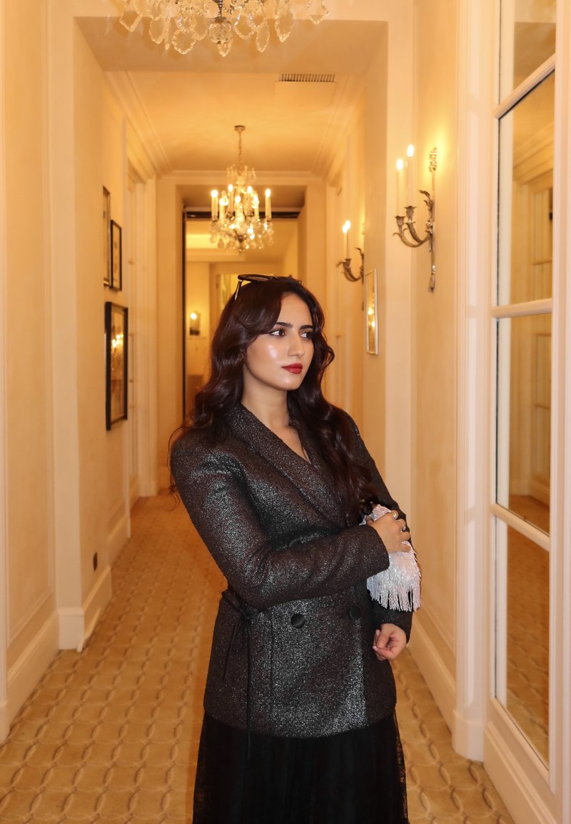 Blazer texturé noir et argent avec une ceinture à la taille, associé à une jupe noire transparente. Situé dans un couloir bien éclairé avec des lustres.