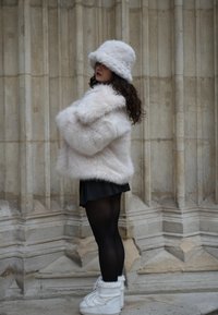 Kosmat, svetlo roza umetno krzno plašč združen z usklajeno kapo v obliki vedra. Črna plisirana krilo in bele zimske škornje do kolen zaokrožijo videz.