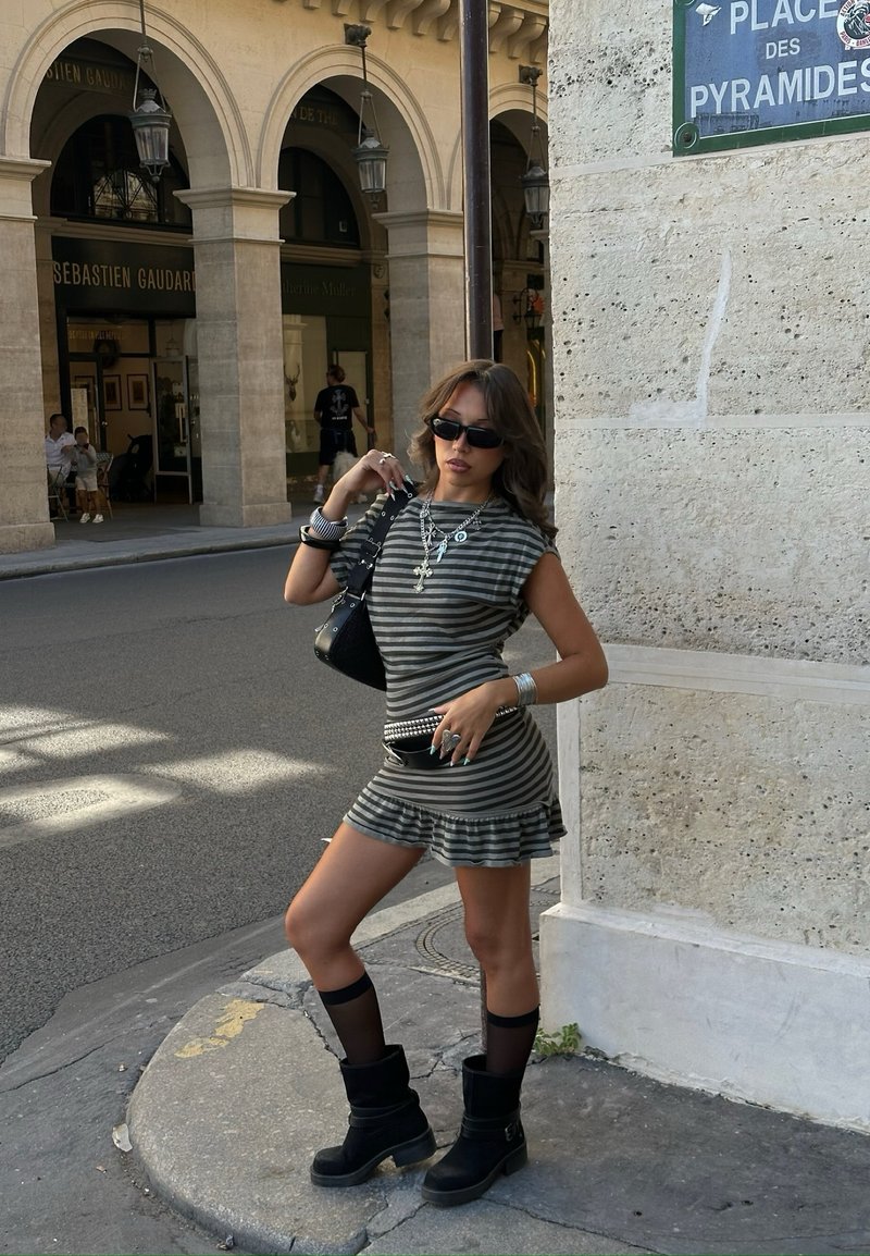 Femme en robe rayée, bottes noires et lunettes de soleil pose à un coin de rue près d'un panneau Place des Pyramides avec un bâtiment voûté en arrière-plan.