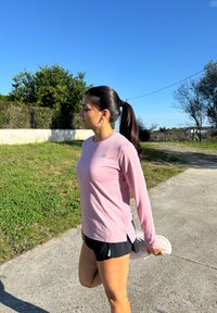 Top atletico rosa chiaro a maniche lunghe con spacchi laterali, abbinato a pantaloni corti da corsa neri plissettati. Ambientazione all'aperto con erba e cielo blu limpido.
