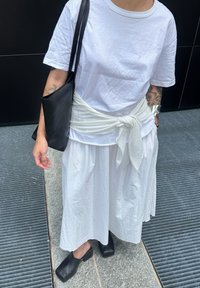 Witte t-shirt, witte rok met een strikband, en zwarte blokhak schoenen. Zwarte schoudertas. Huid met zichtbare tatoeages.